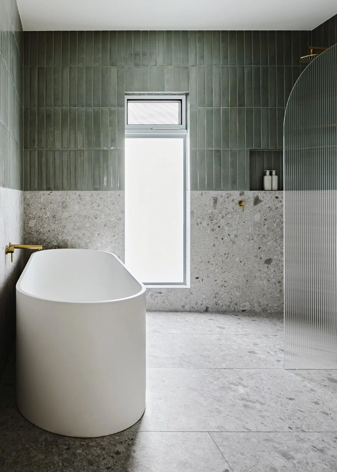Freestanding bath in a modern wet room bathroom with terrazzo tiles, vertical green wall tiles, and a curved fluted glass shower screen